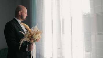 Groom Holding Wedding Bouquet, Thoughtful groom in a black suit holding a rustic wedding bouquet, gazing through a bright window. video