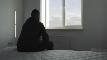 soledad en el dormitorio, hombre sentado en cama frente a un ventana en un minimalista dormitorio, reflejando o contemplando video