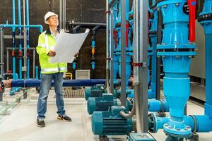 asian man worker in protective uniform and with hardhat using tablet for checking temperature in pipes Factory interior. photo