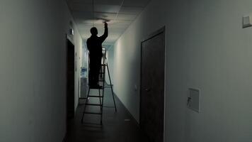 silhouet een meester elektricien is staand Aan een ladder en veranderen een licht lamp in een donker gang video