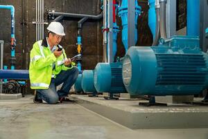 asian man worker in protective uniform and with hardhat using tablet for checking temperature in pipes Factory interior. photo