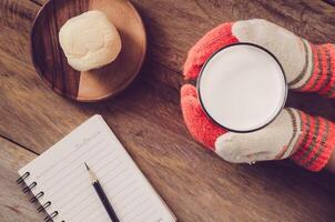 Hand glove holding a glass of warm milk at breakfast and notebook pencil on wood floor photo