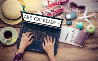 Hands on the keyboard of a notebook's screen displays, wo ARE YOU READY and travel accessories are placed on a wooden table. The concept plan tour photo
