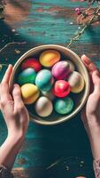 AI generated Hands holding a bowl filled with decorated Easter eggs, set against a green textured backdrop. Top view. Vertical format. Ideal For event planning, festive articles, and home photo