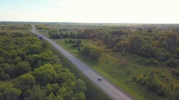 Aerial view of Russian country road and moving cars in summer season surrounded by green trees and fields against blue sky. Clip. Beautiful summer landscape video