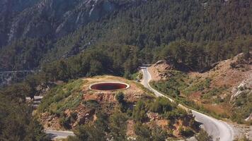 antenne visie van de klein kleurrijk vijver met rood water en lucht reflectie in de buurt de serpentijn en bergen gedekt met groen Woud Aan de achtergrond. klem. mooi zomer landschap video