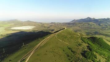 Beautiful view of path on the top of high green hill near the meadows and lake with mountain chain on the background against blue sky. Shot. Aerial view video