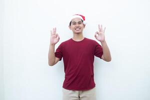 Young Asian man wearing a Santa Claus hat with a hand making the ok gesture isolated by a white background for visual communication photo