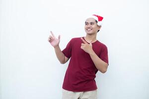 joven asiático hombre vistiendo un Papa Noel claus sombrero sonriente, conmoción y señalando a su lado aislado por blanco antecedentes para visual comunicación foto