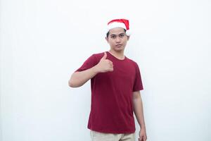 joven asiático hombre vistiendo un Papa Noel claus sombrero sonriente, conmoción y señalando a su lado aislado por blanco antecedentes para visual comunicación foto