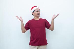 joven asiático hombre vistiendo un Papa Noel claus sombrero sonriente, conmoción y señalando a su lado aislado por blanco antecedentes para visual comunicación foto