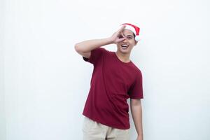 joven asiático hombre vistiendo un Papa Noel claus sombrero con un mano haciendo el Okay gesto aislado por un blanco antecedentes para visual comunicación foto