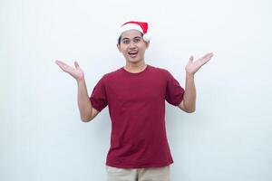 joven asiático hombre vistiendo un Papa Noel claus sombrero sonriente, conmoción y señalando a su lado aislado por blanco antecedentes para visual comunicación foto