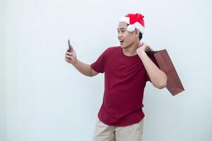 joven asiático hombre vistiendo un Papa Noel claus sombrero participación un teléfono inteligente y un compras bolso con expresiones de sonrisa, choque, y sorpresa, aislado en contra un blanco antecedentes para visual comunicación foto