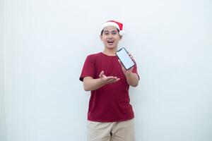 Young Asian man wearing a Santa Claus hat holding a smartphone, and pointing to the phone with a smile, look of shock, and surprise isolated against a white background for visual communication photo