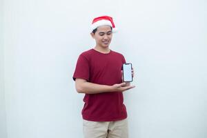 Young Asian man wearing a Santa Claus hat holding a smartphone, and pointing to the phone with a smile, look of shock, and surprise isolated against a white background for visual communication photo
