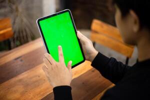 Mock up photo of a close up shot featuring a mans hand holding an iPad tablet with a green screen against the background of a wood cafe table