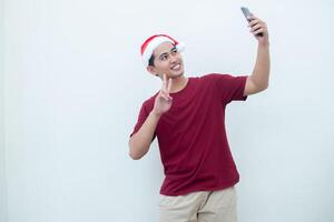 joven asiático hombre vistiendo un Papa Noel claus sombrero, participación un teléfono inteligente y tomando un selfie aislado en contra un blanco antecedentes para visual comunicación foto