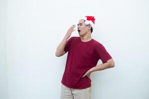 joven asiático hombre vistiendo un Papa Noel claus sombrero expresando cansancio y bostezando aislado por un blanco antecedentes para visual comunicación foto