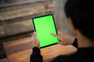 Mock up photo of a close up shot featuring a mans hand holding an iPad tablet with a green screen against the background of a wood cafe table