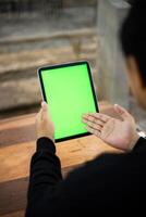 Mock up photo of a close up shot featuring a mans hand holding an iPad tablet with a green screen against the background of a wood cafe table