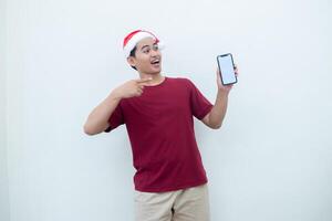 joven asiático hombre vistiendo un Papa Noel claus sombrero participación un teléfono inteligente, y señalando a el teléfono con un sonrisa, Mira de choque, y sorpresa aislado en contra un blanco antecedentes para visual comunicación foto