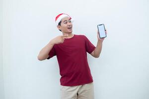 Young Asian man wearing a Santa Claus hat holding a smartphone, and pointing to the phone with a smile, look of shock, and surprise isolated against a white background for visual communication photo