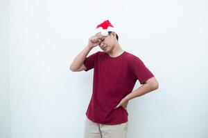 joven asiático hombre vistiendo un Papa Noel claus sombrero expresando un dolor de cabeza por participación su cabeza y demostración señales de dolor aislado por un blanco antecedentes para visual comunicación foto