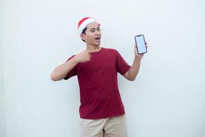 joven asiático hombre vistiendo un Papa Noel claus sombrero participación un teléfono inteligente, y señalando a el teléfono con un sonrisa, Mira de choque, y sorpresa aislado en contra un blanco antecedentes para visual comunicación foto