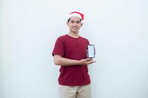 Young Asian man wearing a Santa Claus hat holding a smartphone, and pointing to the phone with a smile, look of shock, and surprise isolated against a white background for visual communication photo