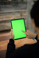 Mock up photo of a close up shot featuring a mans hand holding an iPad tablet with a green screen against the background of a wood cafe table