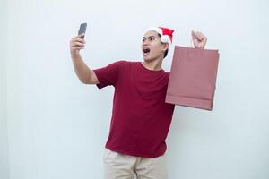joven asiático hombre vistiendo un Papa Noel claus sombrero participación un teléfono inteligente y un compras bolso con expresiones de sonrisa, choque, y sorpresa, aislado en contra un blanco antecedentes para visual comunicación foto