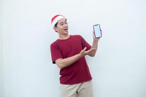 Young Asian man wearing a Santa Claus hat holding a smartphone, and pointing to the phone with a smile, look of shock, and surprise isolated against a white background for visual communication photo