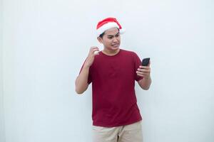 joven asiático hombre vistiendo un Papa Noel claus sombrero participación un teléfono inteligente y expresando un sonrisa, conmoción y sorpresa aislado por un blanco antecedentes para visual comunicación foto