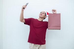 joven asiático hombre vistiendo un Papa Noel claus sombrero participación un teléfono inteligente y un compras bolso con expresiones de sonrisa, choque, y sorpresa, aislado en contra un blanco antecedentes para visual comunicación foto