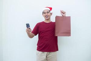 joven asiático hombre vistiendo un Papa Noel claus sombrero participación un teléfono inteligente y un compras bolso con expresiones de sonrisa, choque, y sorpresa, aislado en contra un blanco antecedentes para visual comunicación foto