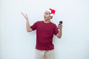 Young Asian man wearing a Santa Claus hat holding a smartphone and expressing a smile, surprise, and pointing to the side, isolated by a white background for visual communication photo