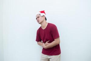 Young Asian man wearing a Santa Claus hat expressing stomachache by holding his stomach in pain isolated by a white background for visual communication photo