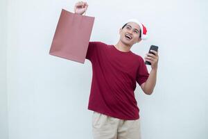 joven asiático hombre vistiendo un Papa Noel claus sombrero participación un teléfono inteligente y un compras bolso con expresiones de sonrisa, choque, y sorpresa, aislado en contra un blanco antecedentes para visual comunicación foto