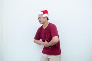 joven asiático hombre vistiendo un Papa Noel claus sombrero expresando dolor de estómago por participación su estómago en dolor aislado por un blanco antecedentes para visual comunicación foto