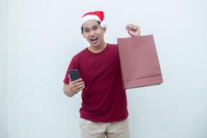 joven asiático hombre vistiendo un Papa Noel claus sombrero participación un teléfono inteligente y un compras bolso con expresiones de sonrisa, choque, y sorpresa, aislado en contra un blanco antecedentes para visual comunicación foto