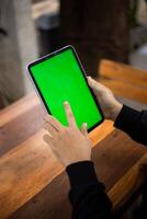 Mock up photo of a close up shot featuring a mans hand holding an iPad tablet with a green screen against the background of a wood cafe table