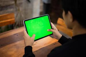 Mock up photo of a close up shot featuring a mans hand holding an iPad tablet with a green screen against the background of a wood cafe table