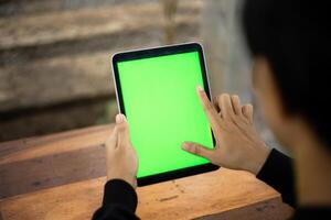 burlarse de arriba foto de un cerca arriba Disparo presentando un mans mano participación un ipad tableta con un verde pantalla en contra el antecedentes de un madera café mesa