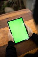 Mock up photo of a close up shot featuring a mans hand holding an iPad tablet with a green screen against the background of a wood cafe table