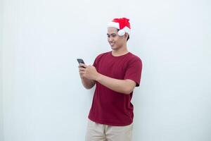 joven asiático hombre vistiendo un Papa Noel claus sombrero participación un teléfono inteligente y expresando un sonrisa, conmoción y sorpresa aislado por un blanco antecedentes para visual comunicación foto