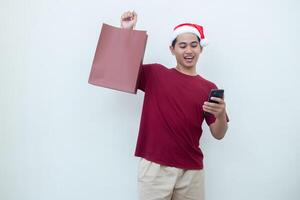 joven asiático hombre vistiendo un Papa Noel claus sombrero participación un teléfono inteligente y un compras bolso con expresiones de sonrisa, choque, y sorpresa, aislado en contra un blanco antecedentes para visual comunicación foto