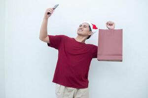 joven asiático hombre vistiendo un Papa Noel claus sombrero participación un teléfono inteligente y un compras bolso con expresiones de sonrisa, choque, y sorpresa, aislado en contra un blanco antecedentes para visual comunicación foto