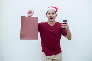 joven asiático hombre vistiendo un Papa Noel claus sombrero participación un teléfono inteligente y un compras bolso con expresiones de sonrisa, choque, y sorpresa, aislado en contra un blanco antecedentes para visual comunicación foto