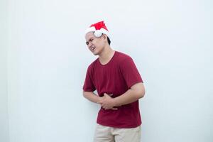 Young Asian man wearing a Santa Claus hat expressing stomachache by holding his stomach in pain isolated by a white background for visual communication photo